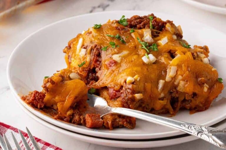 A serving of chili dog casserole plated in a white bowl, topped with melted cheese, diced onions, and garnished with parsley, with a fork placed beside it on the plate