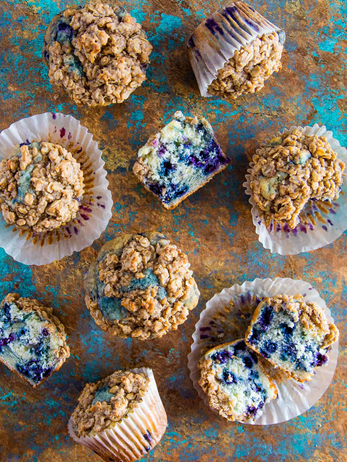 A mix of whole and cut muffins scattered on a rustic blue surface, showing the soft interior and juicy blueberries.